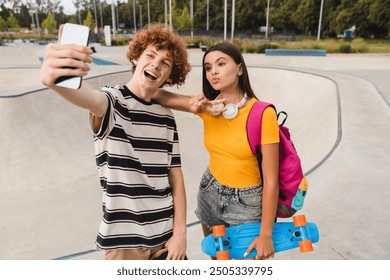 Couple of teenagers classmates students high school pupils friends boy girl bloggers in casual with skateboards holding smart mobile phone taking selfie shot having fun standing against skate park - Powered by Shutterstock