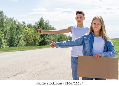 A couple of teenagers are trying to stop a car on the road with an empty cardboard sign, a mock-up. Car travel. Young people are hitchhiking. Two young people tourists hitchhiking on the road. High