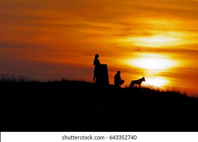 A Couple And Their Dog Silhouetted At Sunset On Painswick Beacon In Summer, The Cotswolds, Gloucestershire, England, UK