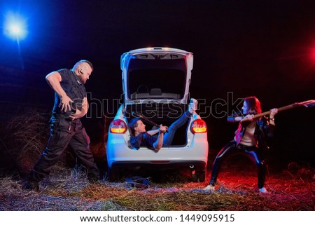 Couple of thugs near car trying to kill young guy in the trunk at night time and colored red and blue light around. Photoshoot about life of gungsters in Russia