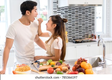 Couple Together In Kitchen Room, Young Asian Woman Holding Vegetables To Man Eating Each For Cooking Dinner Healthy Food Menu At Home Couple Together Romantic 