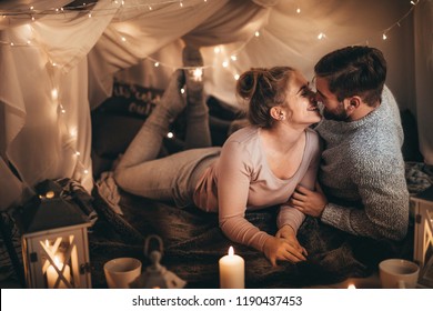 Couple Together On Bed In A Room Decorated With Candle Lights And Tiny Serial Light Bulbs. Smiling Woman Spending Happy Time With Her Husband In Bedroom.