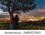 Couple tourist taking a photo from Mount Eden with iconic tree and illuminated city with sky tower at Auckland, New Zealand