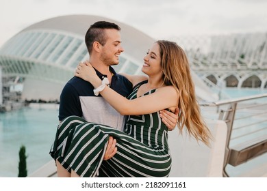 A Couple Of Tourists Are Having Fun In A Modern Urban Space On A Date At The Sunset In Valencia. A Guy Is Holding In His Hands And Kissing His Girlfriend In The Evening In Spain