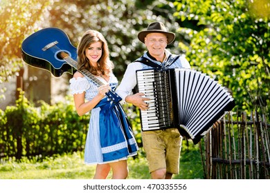 Couple in traditional bavarian clothes standing in the garden in front of wooden fence, playing accordion, holding guitar. Oktoberfest.