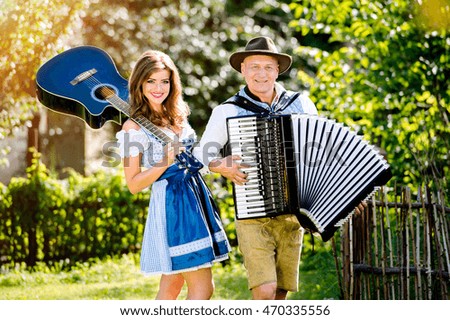 Couple in traditional bavarian clothes standing in the garden in front of wooden fence, playing accordion, holding guitar. Oktoberfest.