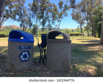 A Couple Of Trash Cans At The Park, With A Folding Chair Next To Them. Maybe It Broke During The Big Baseball Game!