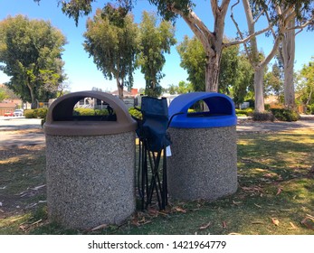 A Couple Of Trash Cans At The Park, With A Folding Chair Next To Them. Maybe It Broke During The Big Baseball Game!