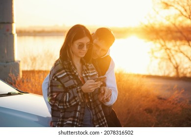 Couple Traveler Stands By Car At Sunset In Countryside. Girl Uses Mobile App Map In Smartphone To Find Out The Way. Tourists Checking Location In Smartphone Map. Soft Focus