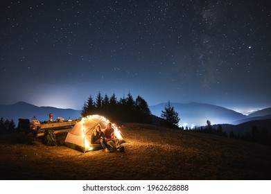 Couple of travelers set up camp on night mountain lawn and relaxing in iluminated tourist tent after dinner. Light of surrounding villages, mountain peaks under evening starry sky on the background.