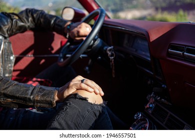 Couple Traveling Together In An Old Red Muscle Car. Cropped Shot Of Man In Leather Jacket With One Hand On Steering Wheel And Other On Girlfriend's Knee. Close Up, Copy Space, Background.