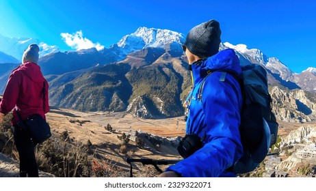 A couple trekking along Annapurna Circuit Trek, Himalayas, Nepal. Snow caped Annapurna chain in the back. They are surrounded with dried grass. Harsh landscape. Adventure and exploration.