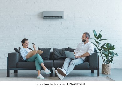 couple turning on air conditioner during the summer heat while sitting on sofa with book and laptop 