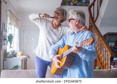 Couple Of Two Happy Seniors Or Mature And Old People Singing And Dancing Together At Home Indoor. Retired Man Playing The Guitar While His Wife Is Singing With A Remote Control Of TV.