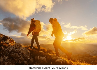 couple of two hikers, man and woman, together with their dog, ascending a mountain peak at sunset with the sun in the background. sport and adventure. Weekend activities.