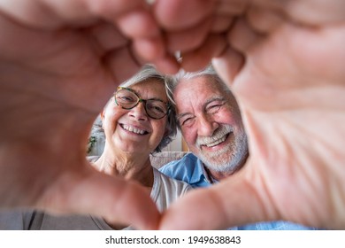 Couple Of Two Old And Happy Seniors Having Fun At Home On The Sofa Doing A Heart Shape With Their Hands And Fingers Looking At The Camera.