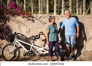 Couple Of Two Seniors And Mature People Standing With The Wall At The Background In A Park With Their Tandem Or Doble Bike Smiling And Talking