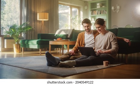 Couple Use Laptop Computer, while Sitting on the Living Floor room of their Apartment. Boyfriend and Girlfriend Talk, Shop on Internet, Choose Product to Order Online, Watch Streaming Service