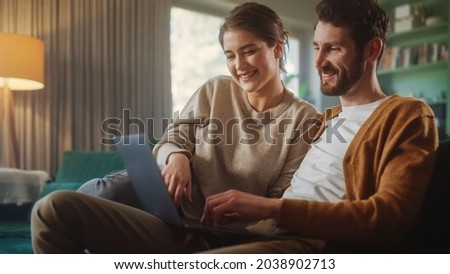 Couple Use Laptop Computer, while Sitting on a Couch in the Apartment. Boyfriend and Girlfriend Talk, Shop on Internet, Choose Product to Order Online, Watch Funny Videos on Streaming Service