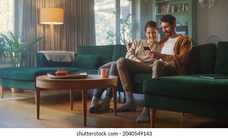 Couple Use Smartphone Device, while Sitting on a Couch in the Cozy Apartment. Boyfriend and Girlfriend Talk, do e-Shopping on Internet, Watching Funny Videos, Use Social Media, Streaming Service. - Shutterstock ID 2040549143