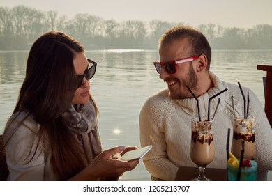 Couple using cellphone while drinking coffee outdoors.