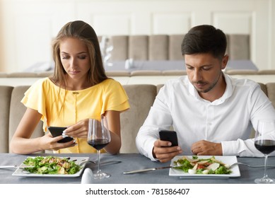 Couple Using Phones On Dinner In Restaurant. Young Man And Woman Looking On Mobile Phones Feeling Bored On Boring Date In Restaurant. Communication Problems. High Quality Image.