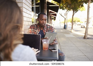 Couple Using Technology At A Table Outside A Cafe