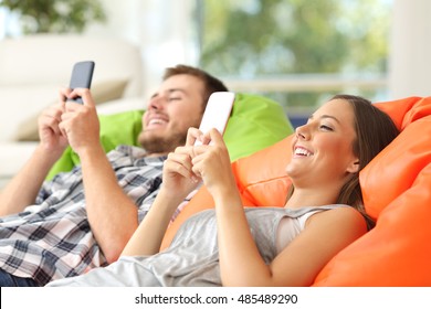Couple using their smart phones lying on a green and orange poufs in the living room at home