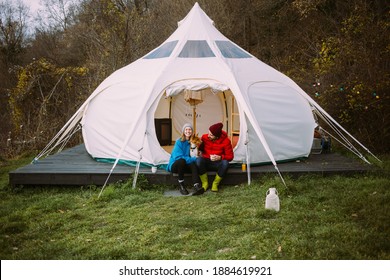 Couple Of Vacation Goers Campers Sit In Front Of Big Glamping Tent. Happy Couple On Romantic Weekend Getaway With Pet Dog. Fun And Luxury Camping Experience In Nature