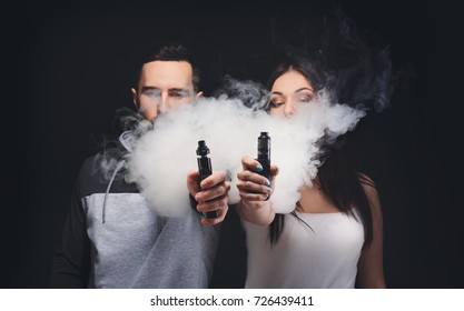 Couple vaping. Unrecognizable young man and woman in the clouds of smoke showing their vapes to camera at black studio background. Relationship and vape addiction concept with copy space
