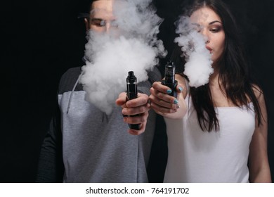 Couple vaping. Unrecognizable young man and woman in the clouds of smoke showing their vapes to camera at black studio background. Relationship and vape addiction concept with copy space