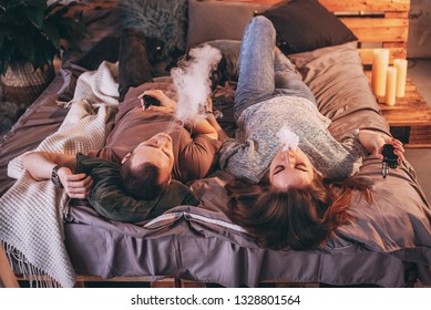 Couple vaping. Young man and woman blowing smoke while sitting on a wooden bed. The concept of relationships and vape dependence with copy space