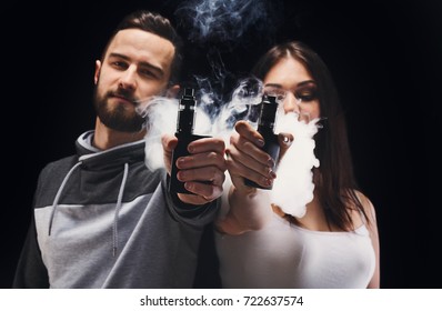 Couple Vaping. Young Man And Woman Blowing Smoke To Join It In One Cloud And Showing Their Vapes To Camera At Black Studio Background. Relationship And Vape Addiction Concept With Copy Space