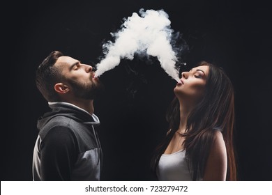 Couple vaping. Young man and woman blowing smoke to join it in one cloud at black studio background. Relationship and vape addiction concept with copy space