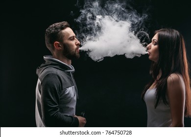 Couple vaping. Young man and woman blowing smoke to join it in one cloud at black studio background. Relationship and vape addiction concept with copy space