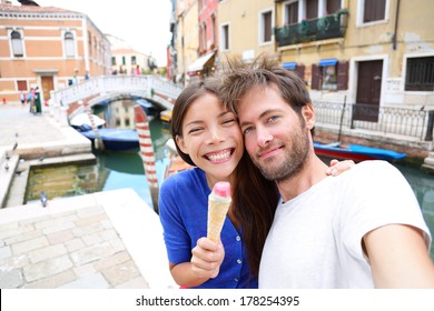 Couple in Venice, eating Ice cream taking selfie self-portrait photo on vacation travel in Italy. Smiling happy Asian woman and Caucasian man in love having fun eating italian gelato food outdoors.
