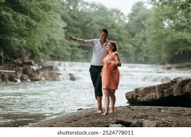 A couple is walking along a riverbank, with the man pointing to something in the distance. The woman is wearing an orange dress and she is enjoying the moment. Concept of romance and adventure - Powered by Shutterstock