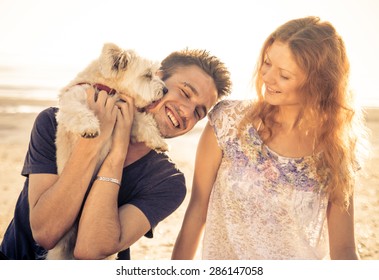 Couple Walking With Dog Outdoor. Man Holding Up The Cute Dog While He Licks His Face