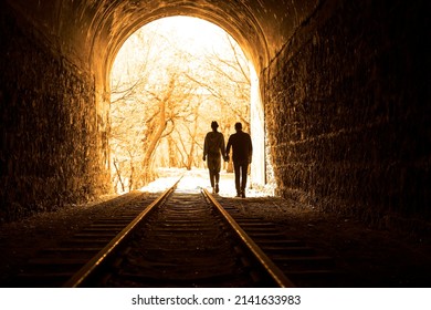 Couple walking hand in hand along the track through a railway tunnel towards the bright light at the other end, they appear as silhouettes against the light. back view. - Powered by Shutterstock