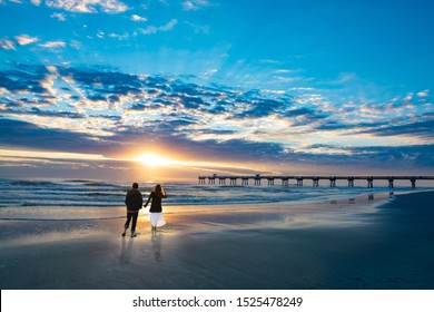 Couple Walking On Beach At Sunrise, Enjoying Time Together. Beautiful Cloudy Sky Over Pier And Sun Reflected On The Beach. People Relaxing On Summer  Vacation. Jacksonville, Florida, USA.