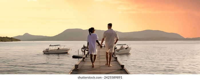 couple walking on a wooden pier in the ocean at sunset in Thailand.Caucasian men and Asian women diverse couple waking at a jetty in the ocean, man and women watching sunset together - Powered by Shutterstock