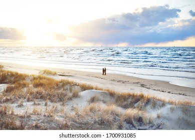 A couple is walking through the Baltic sea shore at sunset. Sand dunes and plants close-up. Colorful evening clouds. Waves and water splashes. Idyllic seascape. Latvia - Powered by Shutterstock