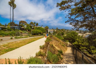 a couple walking wearing colorful clothes standing on a sidewalk surrounded by lush green trees, grass and plants and apartments and homes with blue sky and clouds at Leadbetter Beach in Santa Barbara - Powered by Shutterstock