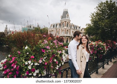 A Couple Walks In Amsterdam. Old City. Love Story. Walk Around The City. Brown-haired Girl In A Beige Trench Coat. The Guy In The Shirt And Jeans. Modern Architecture. Flowers On Bridge Over Canal.