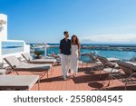 A couple walks on a terrace with lounge chairs, overlooking a marina with docked boats. The setting features Marrakech inspired architecture and a clear blue sky.