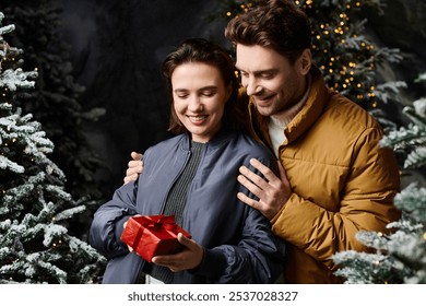 A couple in warm winter jackets smiles as they share a festive present among snow-covered pine trees. - Powered by Shutterstock