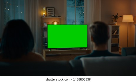 Couple Watches Green Mock-up Screen TV while Sitting on a Couch in the Living Room. Romantic Evening for Boyfriend and Girlfriend.