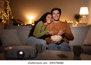 Couple Watches Movie Via Domestic Cinema Projector And Eats Popcorn From Bowl During Evening At Home, Sitting On Sofa In Living Room. Front View, Selective Focus On Happy Spouses