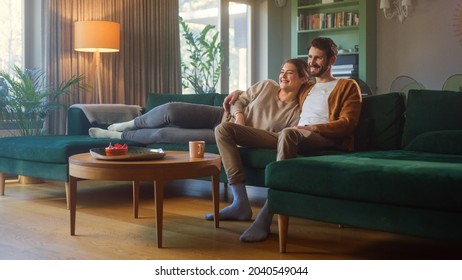 Couple Watches TV Together While Sitting On A Couch In The Living Room. Girlfriend And Boyfriend Embrace, Cuddle, Talk, Smile And Watch Television Streaming Services. Home With Cozy Stylish Interior.