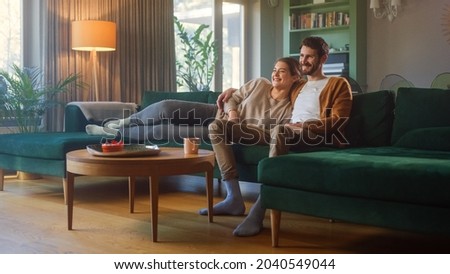 Couple Watches TV together while Sitting on a Couch in the Living Room. Girlfriend and Boyfriend embrace, cuddle, talk, smile and watch Television Streaming Services. Home with Cozy Stylish Interior.